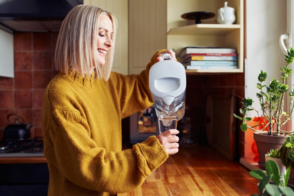 A woman is smiling while pouring filtered water into a glass in the kitchen.